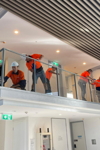 A group of construction workers in orange high-visibility shirts, some wearing hard hats, are working on the railing of an indoor balcony.