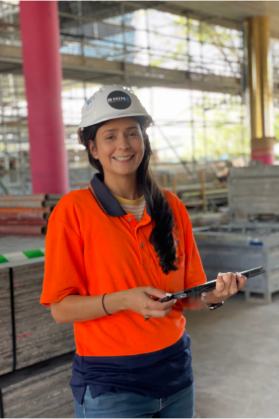 Rhino worker smiling and holding a tool on a construction site, wearing a hard hat and orange shirt.